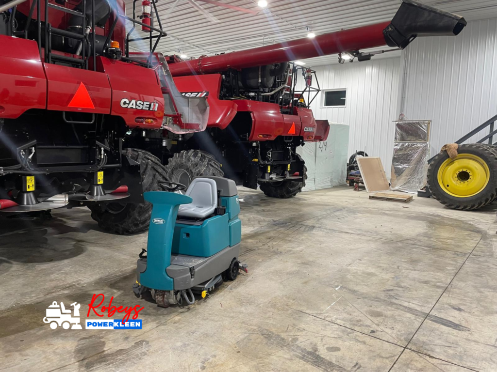 A teal Tennant T7 ride-on floor scrubber from Robey's PowerKleen parked inside a large metal shop building with a massive red Case IH combine harvester in the background.