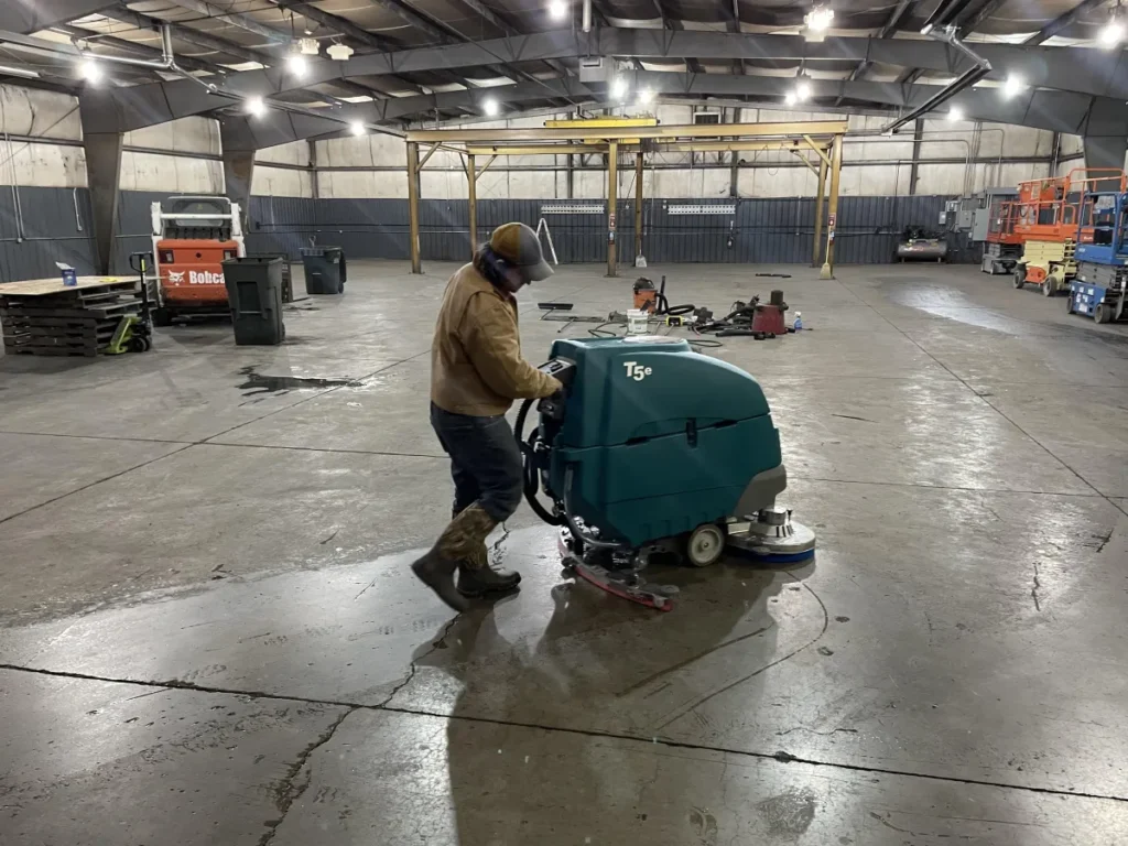 Operator using a Tennant T5e walk-behind floor scrubber inside a large industrial warehouse, cleaning concrete floors in a commercial work environment with equipment and machinery in the background.