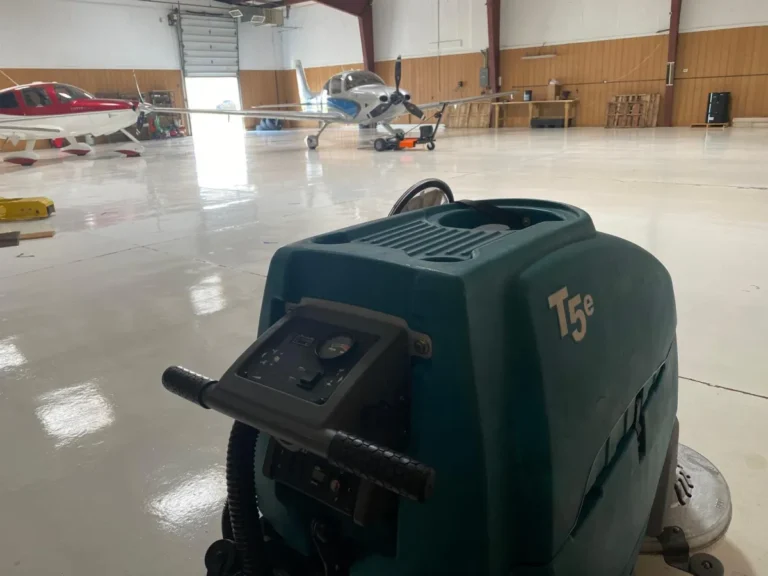 Tennant T5e walk-behind floor scrubber positioned inside an aircraft hangar with polished concrete floors and small airplanes in the background, showcasing commercial floor cleaning equipment for aviation facilities.