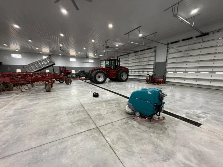 Tennant T5e walk-behind floor scrubber cleaning polished concrete floors inside a large agricultural equipment shop, with tractors, farm machinery, and oversized garage doors in the background.