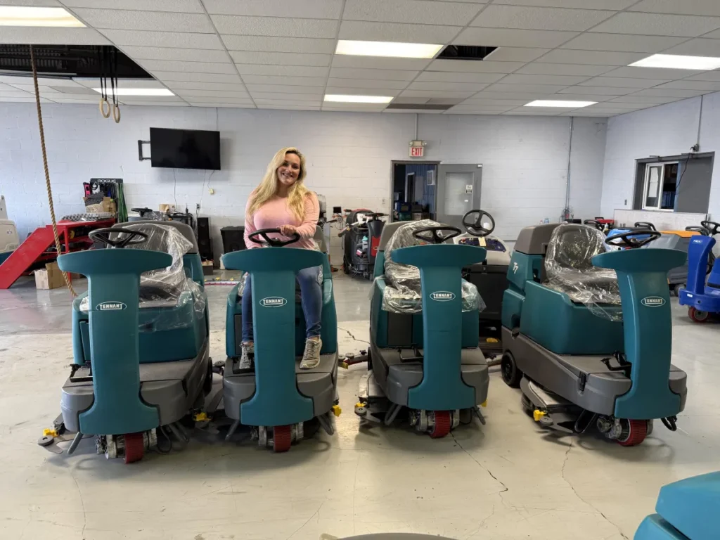Erica sitting in one of the Tennant T7 ride-on floor scrubbers inside the Robey’s PowerKleen warehouse, showcasing available inventory of commercial floor cleaning machines ready for sale or delivery.