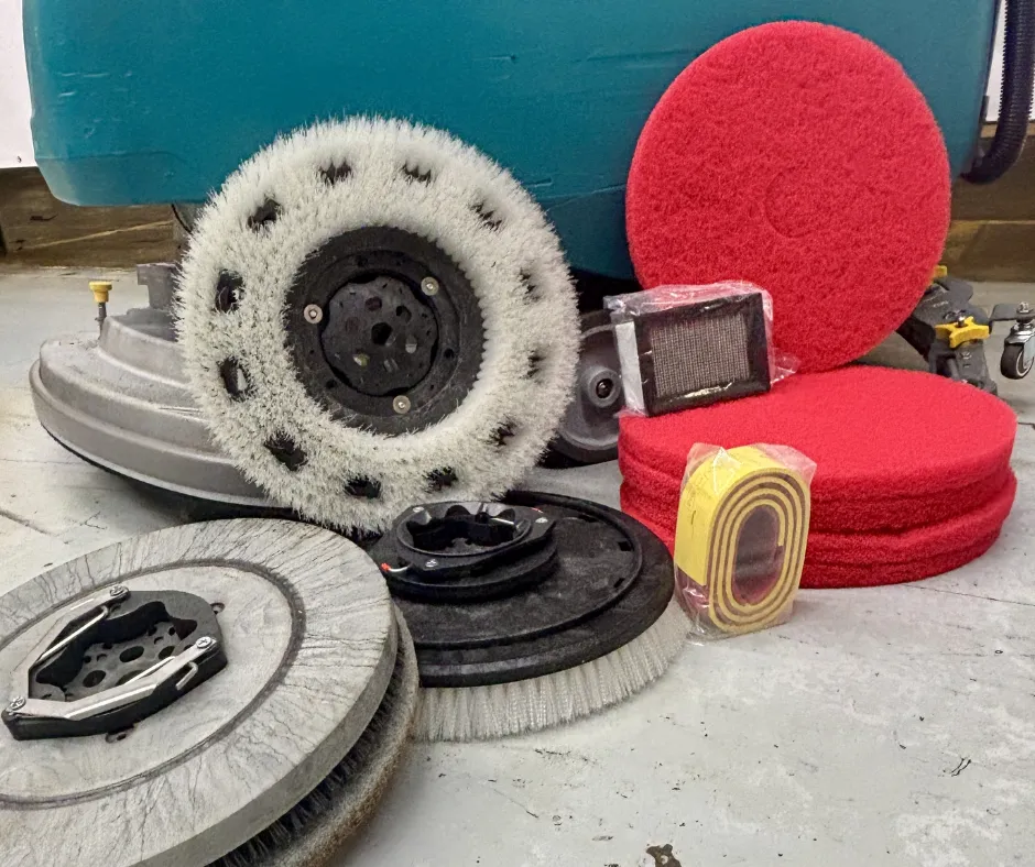 Assorted commercial floor scrubber brushes, cleaning pads, filters, and accessory parts displayed on a concrete floor beside a walk-behind floor scrubber in a warehouse setting.