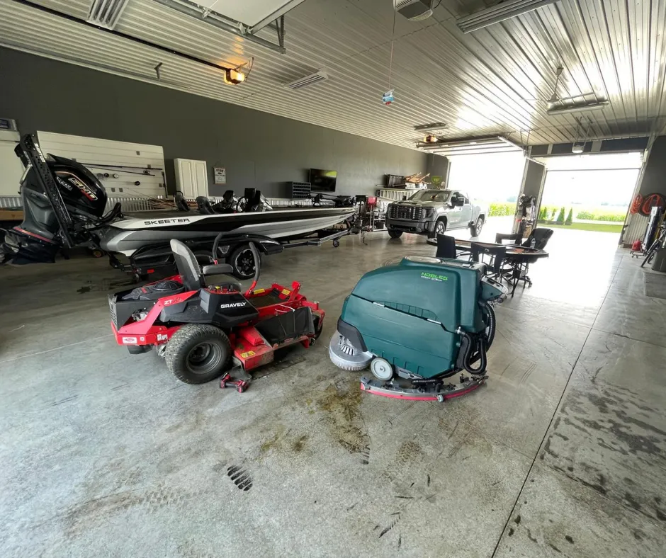 Nobles Speed Scrub SS5 walk-behind floor scrubber parked inside a large commercial garage alongside a zero-turn mower and fishing boat, highlighting its industrial floor cleaning use in equipment storage and workshop environments.