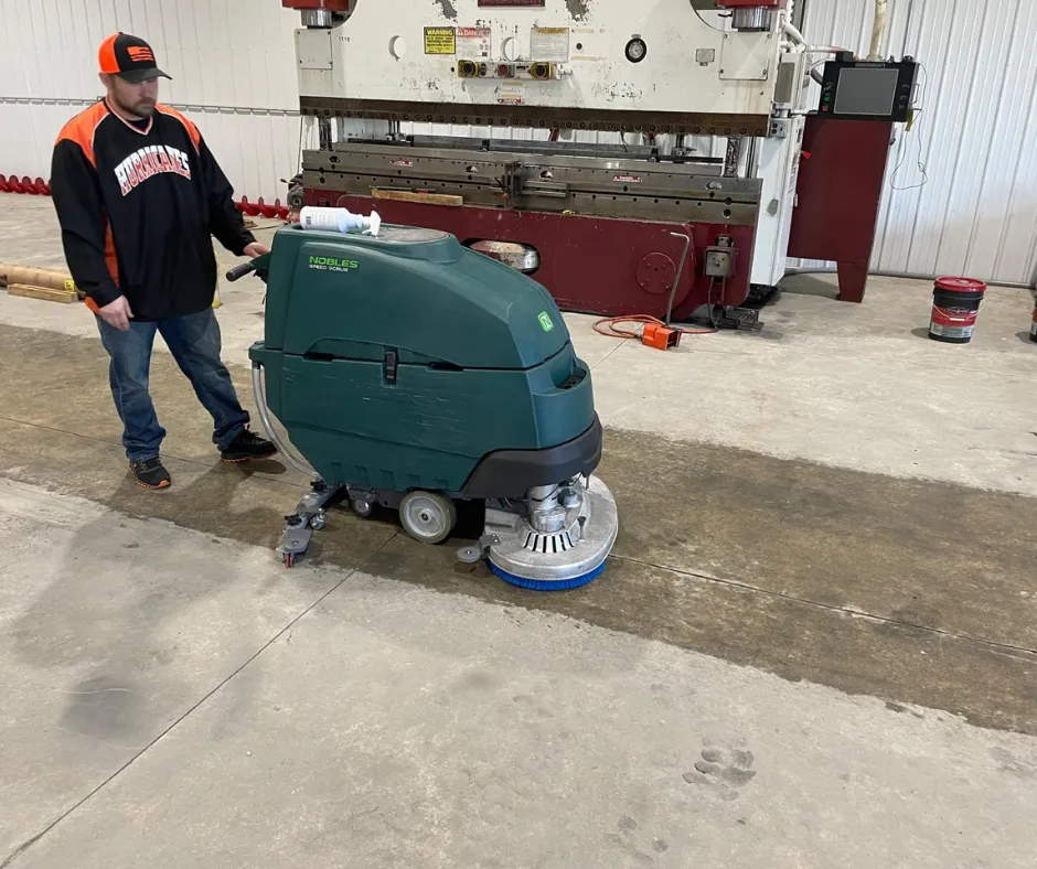 Nobles Speed Scrub SS5 walk-behind floor scrubber being operated on a concrete industrial floor by a worker inside a manufacturing facility, demonstrating commercial floor cleaning performance near heavy machinery.
