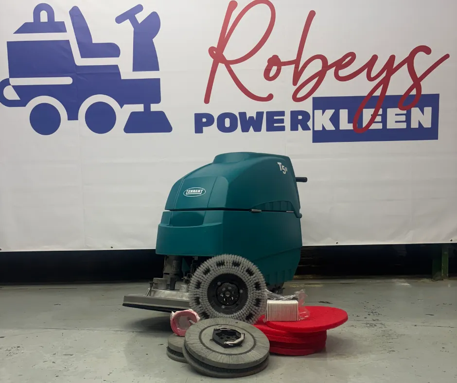 Tennant T5e walk-behind floor scrubber displayed with cleaning pads, brushes, and replacement accessories in front of a Robeys PowerKleen branded backdrop inside a commercial showroom.