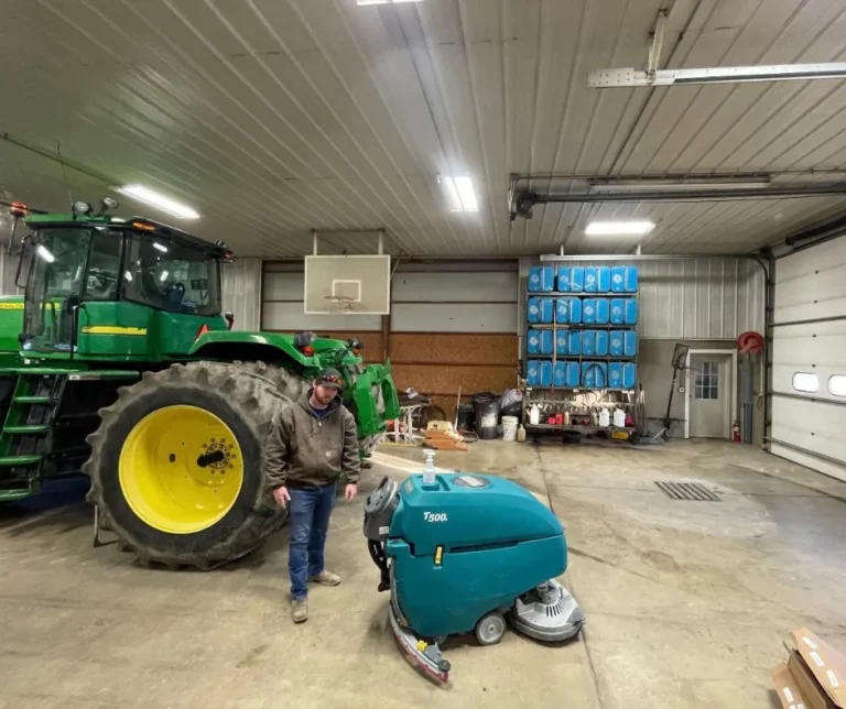 Tennant T500 walk-behind floor scrubber inside a large farm workshop beside a green tractor and an operator standing nearby.