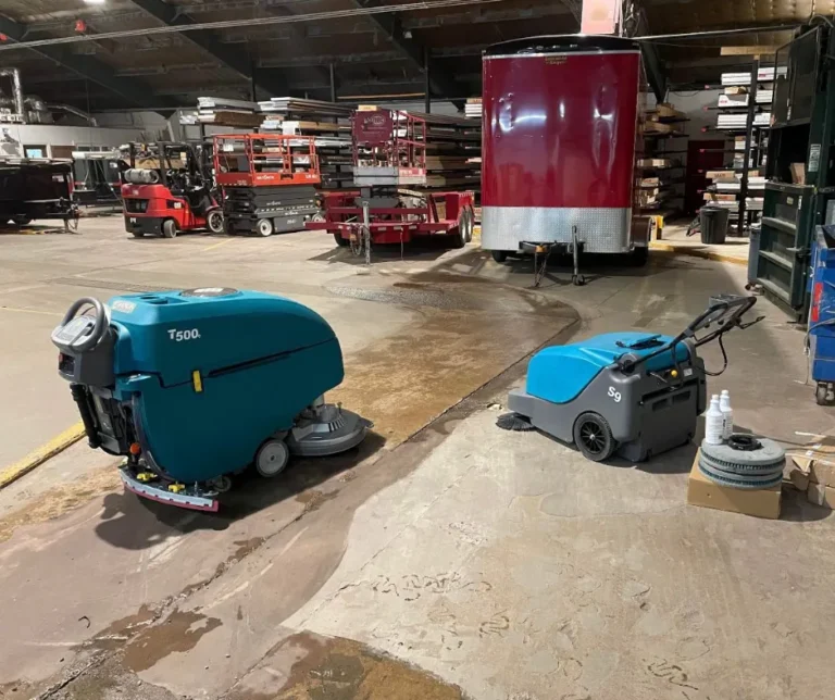 Tennant T500 floor scrubber and S9 sweeper positioned inside an industrial warehouse with trailers, forklifts, and equipment in the background.