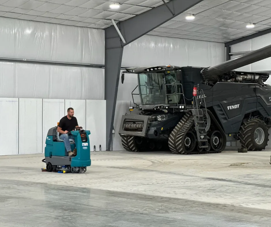 A Tennant T7 AMR ride-on floor scrubber cleaning a large agricultural equipment warehouse, operating near a massive Fendt combine harvester on polished concrete floors.