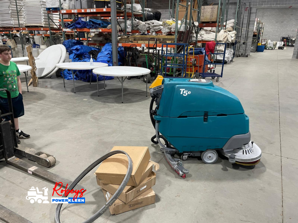 A teal Tennant T5e walk-behind floor scrubber from Robey's PowerKleen parked on a concrete floor inside a large storage facility or warehouse. Folded round tables, blue tarps, and metal storage racks are visible in the background, with some cardboard boxes in the foreground.