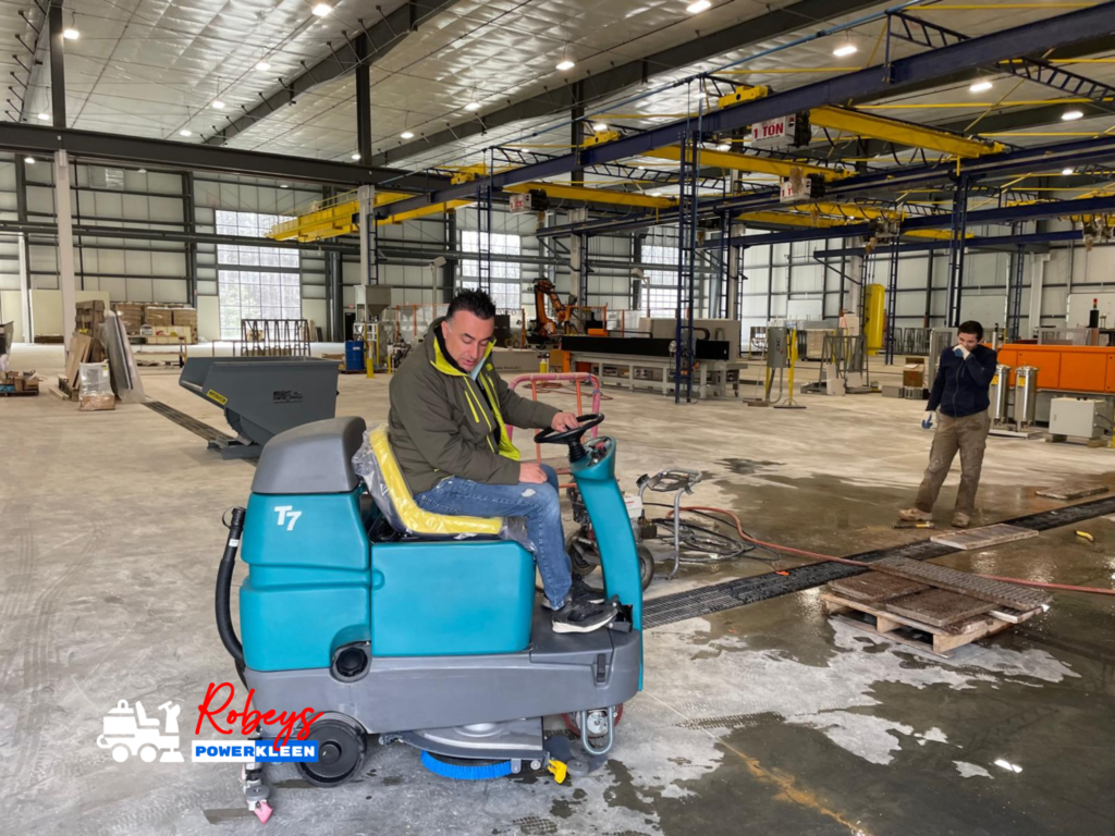 A man operating a teal Tennant T7 ride-on floor scrubber from Robey's PowerKleen on a wet concrete floor inside a large, bright industrial manufacturing facility. Yellow overhead crane beams are visible above, and another worker stands in the background watching.