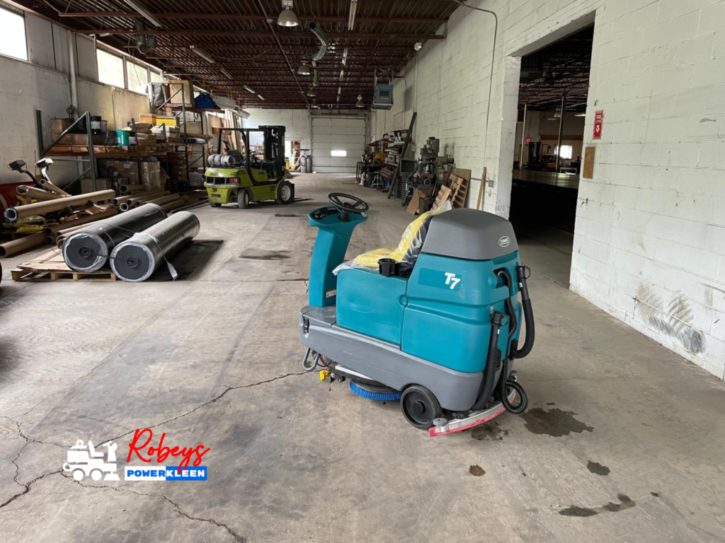 A teal Tennant T7 ride-on floor scrubber from Robey's PowerKleen with protective plastic on its seat, parked on a concrete floor inside a large industrial warehouse. A yellow forklift and metal storage racks are visible in the background.