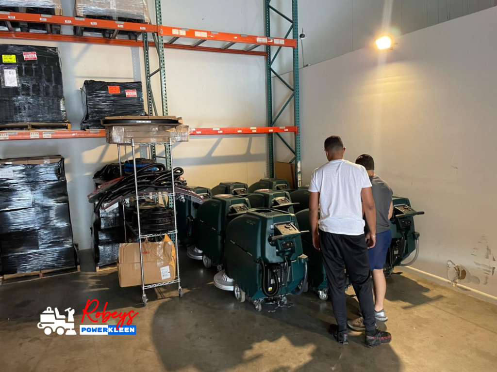 wo men inspecting a lineup of dark green Nobles walk-behind floor scrubbers from Robey's PowerKleen, parked inside a warehouse facility next to tall metal storage racks filled with wrapped pallets. A "Robey's PowerKleen" logo is watermarked in the bottom left corner.