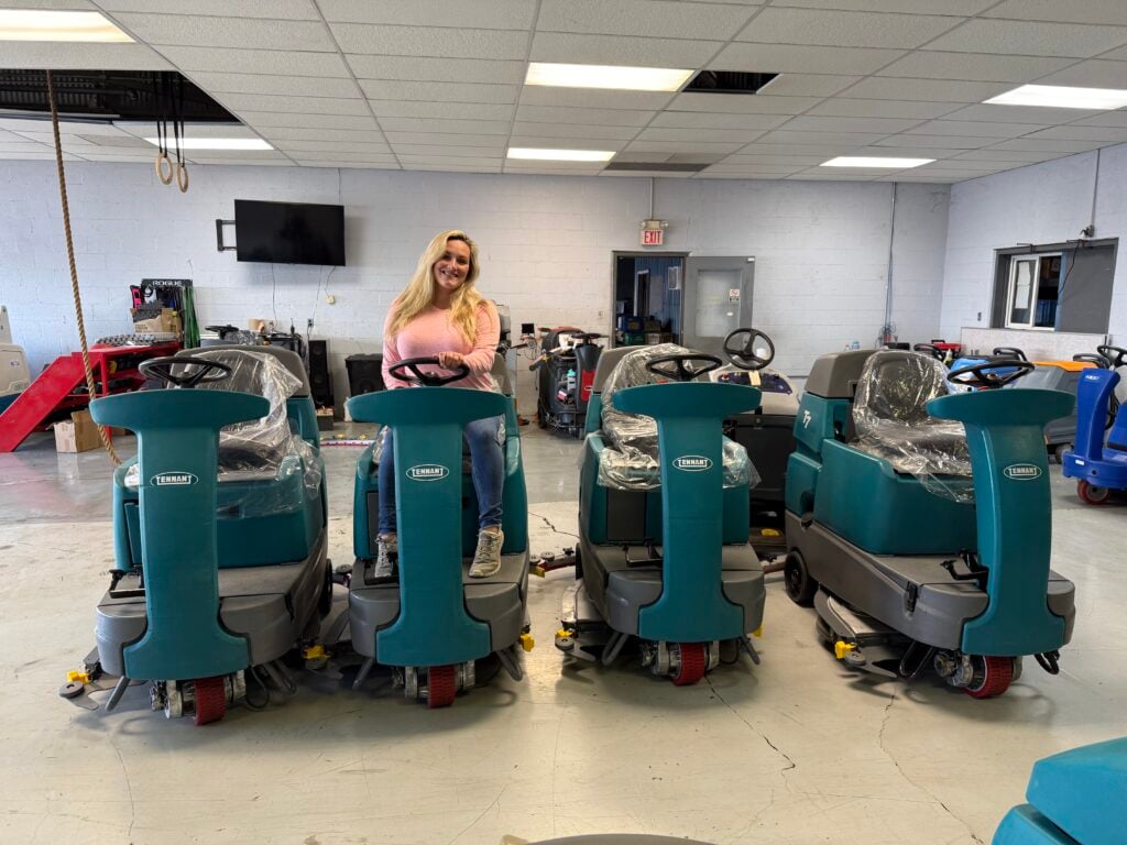 Erica riding on a Tennant T7 ride-on floor scrubber surrounded by multiple Tennant floor scrubbers inside a commercial cleaning equipment showroom.