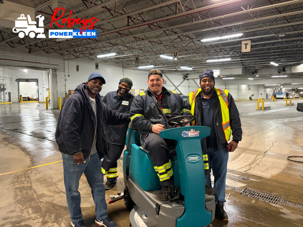 Indianapolis transport team members posing with a Tennant ride-on floor scrubber inside a large industrial facility during a commercial floor cleaning operation.