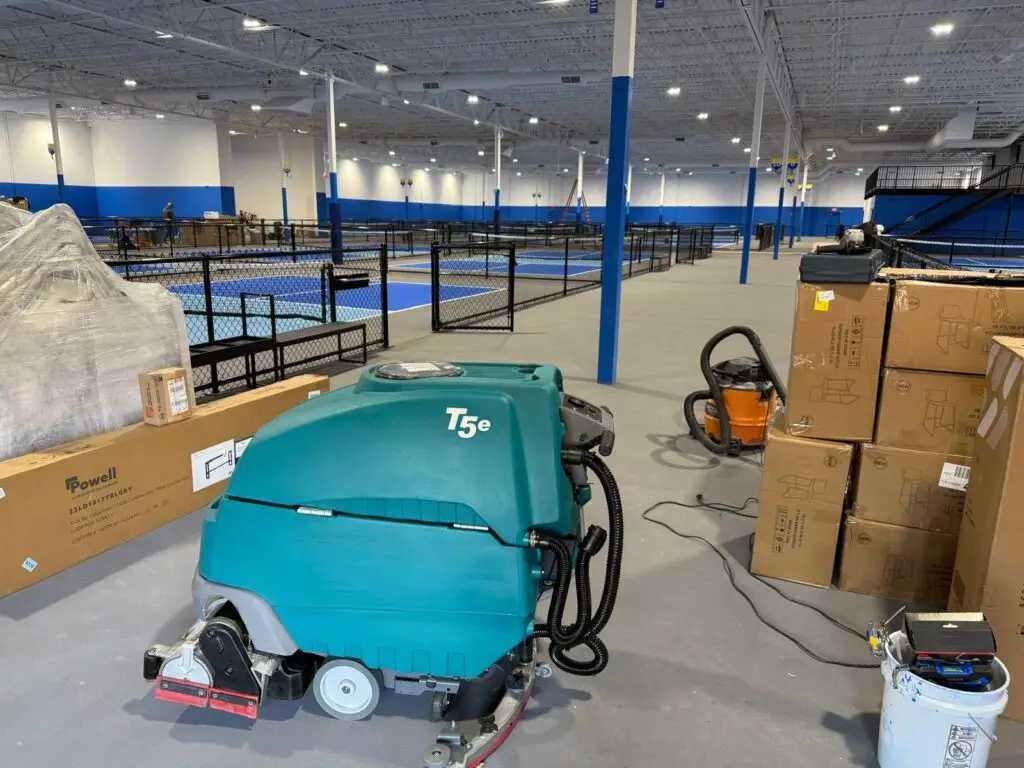 A teal Tennant T5e walk-behind floor scrubber from Robey's PowerKleen parked inside a large indoor sports complex. Cardboard boxes and a shop vacuum sit next to the machine, while several blue indoor pickleball or tennis courts enclosed by black fencing are visible in the background.