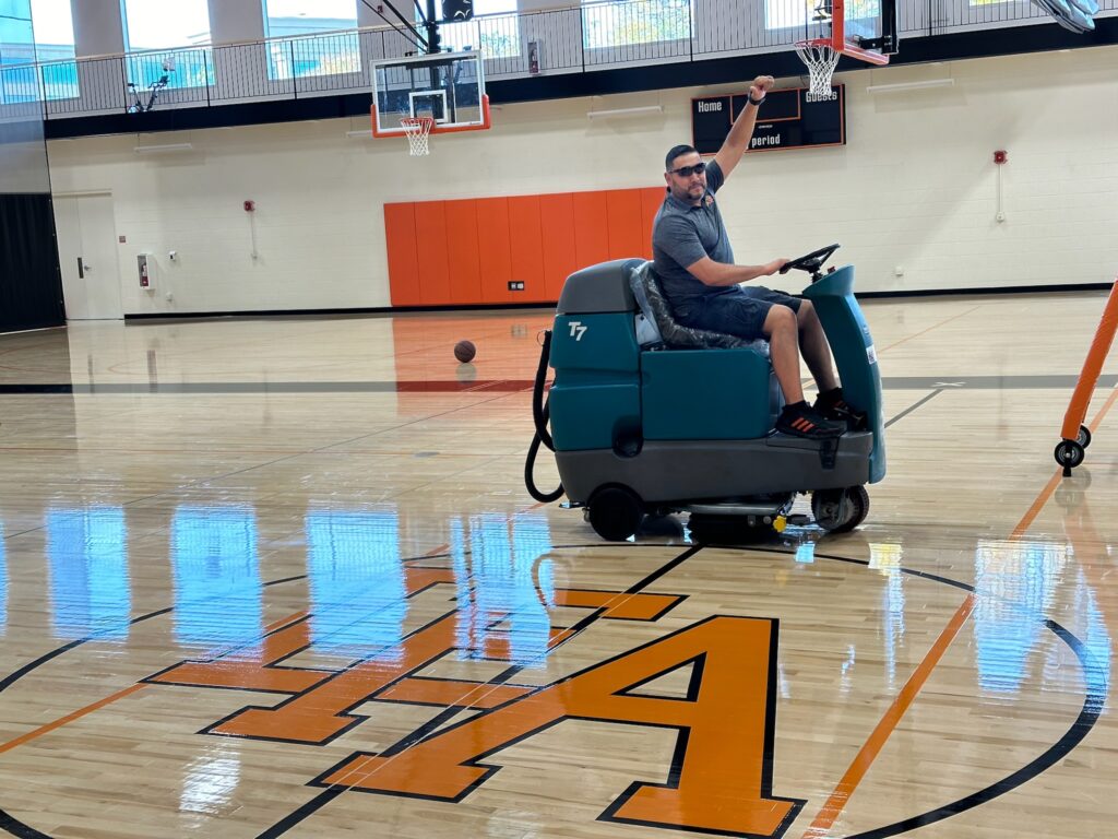 A man happily raises his fist in the air while sitting on a teal Tennant T7 ride-on floor scrubber from Robey's PowerKleen. He is parked on a gleaming hardwood basketball court featuring a large orange "A" logo inside a gymnasium.