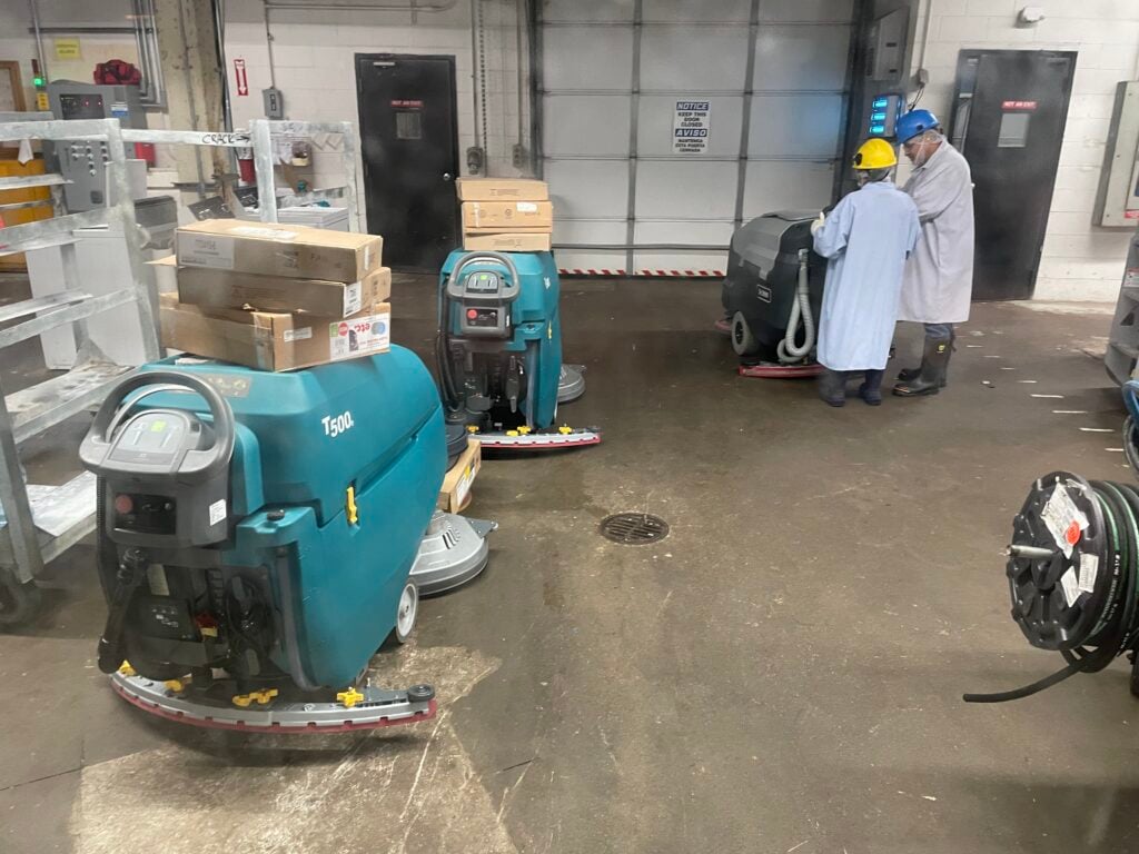 A teal Tennant T500 walk-behind floor scrubber from Robey's PowerKleen parked in the foreground of an industrial facility, with another unit right behind it. In the background, two workers wearing white coats and hard hats are inspecting a third floor scrubber on a wet concrete floor near closed bay doors.