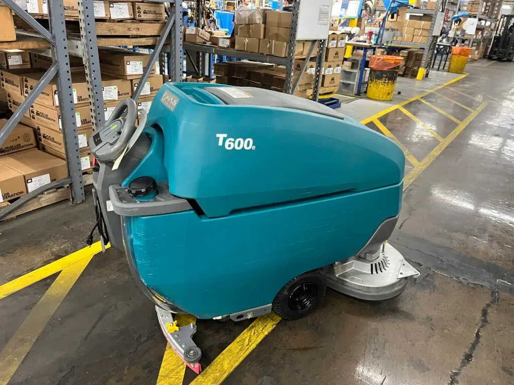 A teal Tennant T600e walk-behind floor scrubber from Robey's PowerKleen parked on a concrete floor with yellow safety lines inside a large industrial warehouse. Tall metal shelving units filled with packaged boxes and supplies are visible in the background.