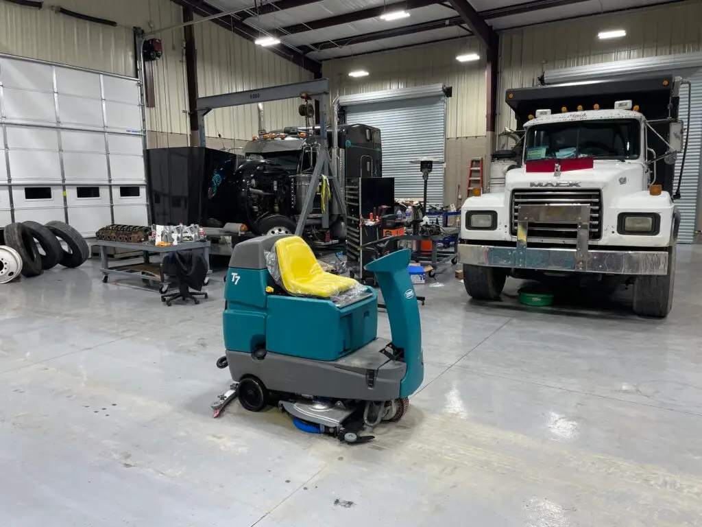 A teal Tennant T7 ride-on floor scrubber with a yellow seat, parked on a clean, light gray concrete floor inside a large diesel mechanic shop. A large white Mack dump truck and a black semi-truck cab are visible in the background, alongside various shop tools, parts, and equipment.