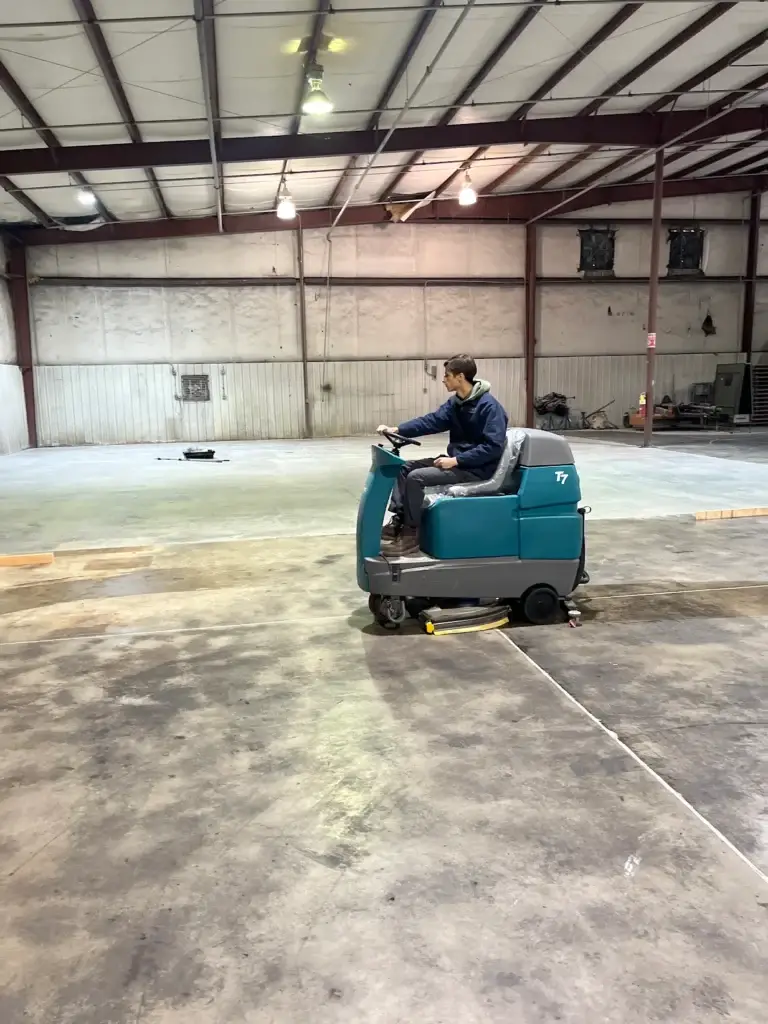 A man operating a teal Tennant T7 ride-on floor scrubber from Robey's PowerKleen across a massive, partially wet concrete floor inside a large, empty industrial warehouse facility.