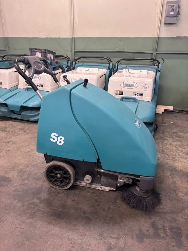 Tennant S8 walk-behind floor sweeper machine inside a warehouse with additional Tennant cleaning equipment in the background.
