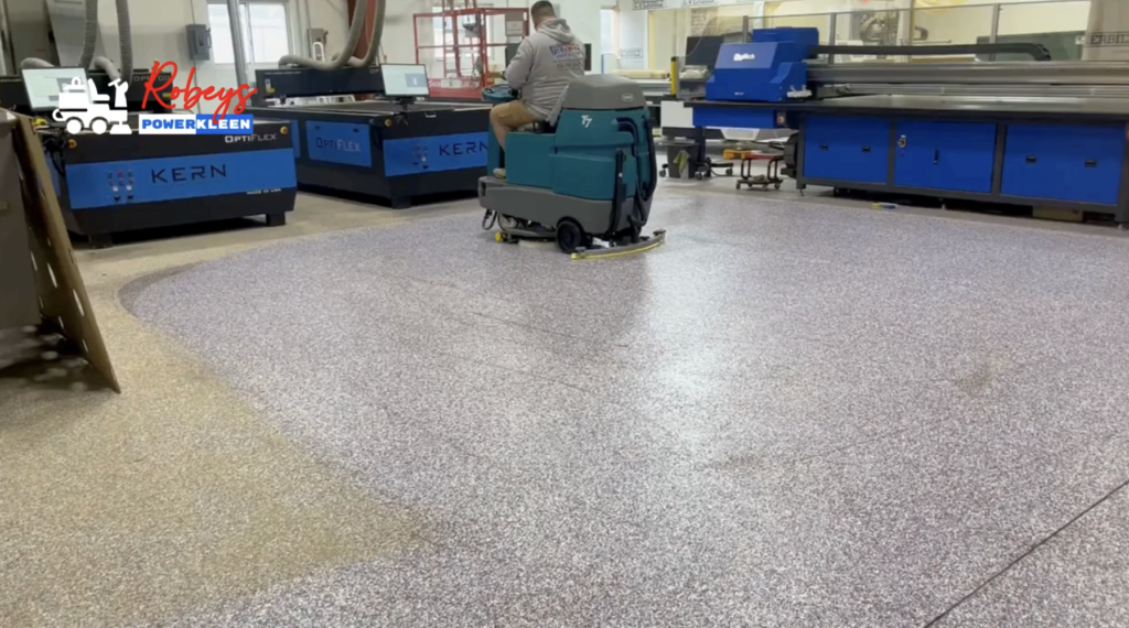 A man operating a teal Tennant T7 ride-on floor scrubber from Robey's PowerKleen on a speckled epoxy floor inside an industrial manufacturing facility. Large blue Kern machinery is visible in the background.