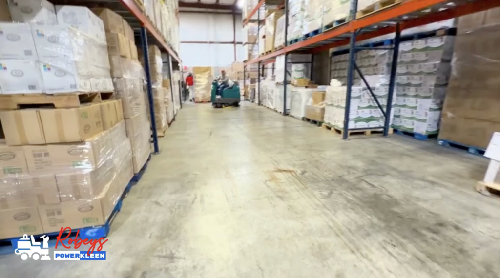 A teal Tennant ride-on floor scrubber from Robey's PowerKleen parked on a concrete floor in the middle of a wide warehouse aisle. Tall metal pallet racks filled with stacked boxes line both sides, and a worker wearing a red high-visibility jacket walks away in the background.