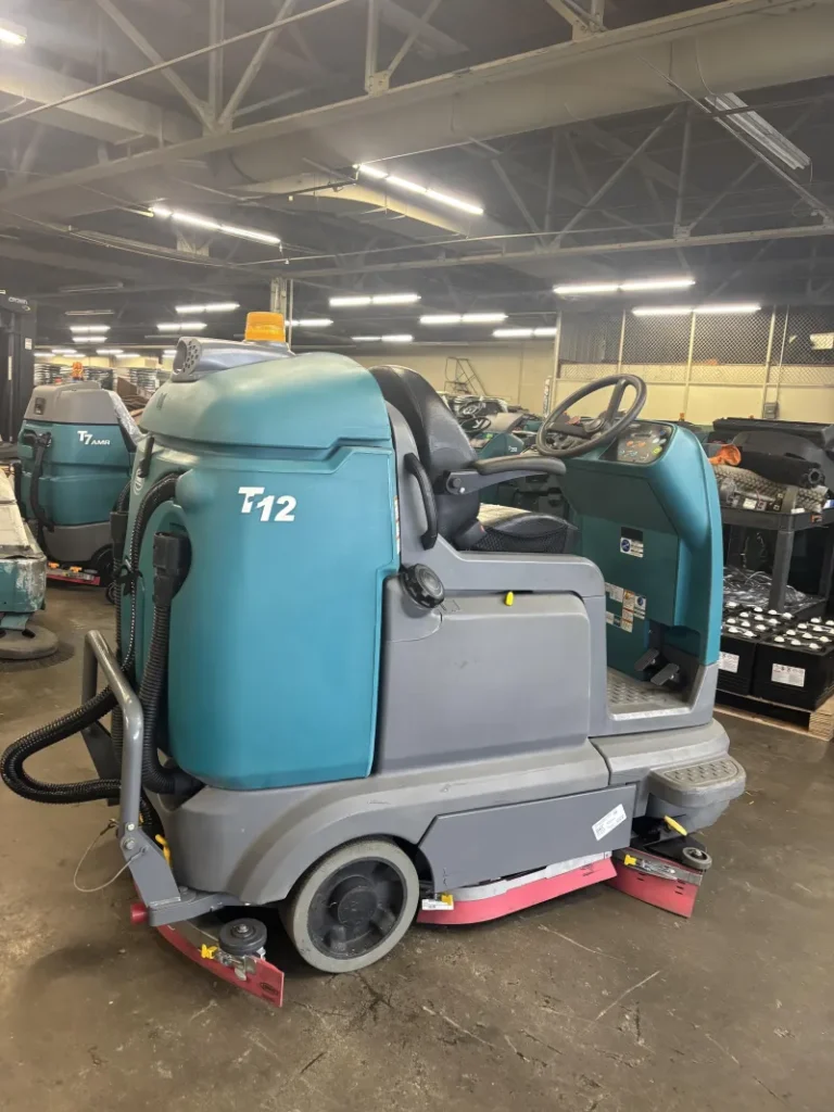 Blue and gray Tennant T12 ride-on floor scrubber machine parked inside an industrial warehouse, featuring an operator seat, steering wheel, and front scrubbing deck for large commercial floor cleaning applications.