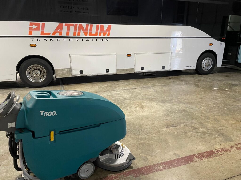 Tennant T500 walk-behind floor scrubber parked inside a transportation facility beside a Platinum Transportation coach bus.