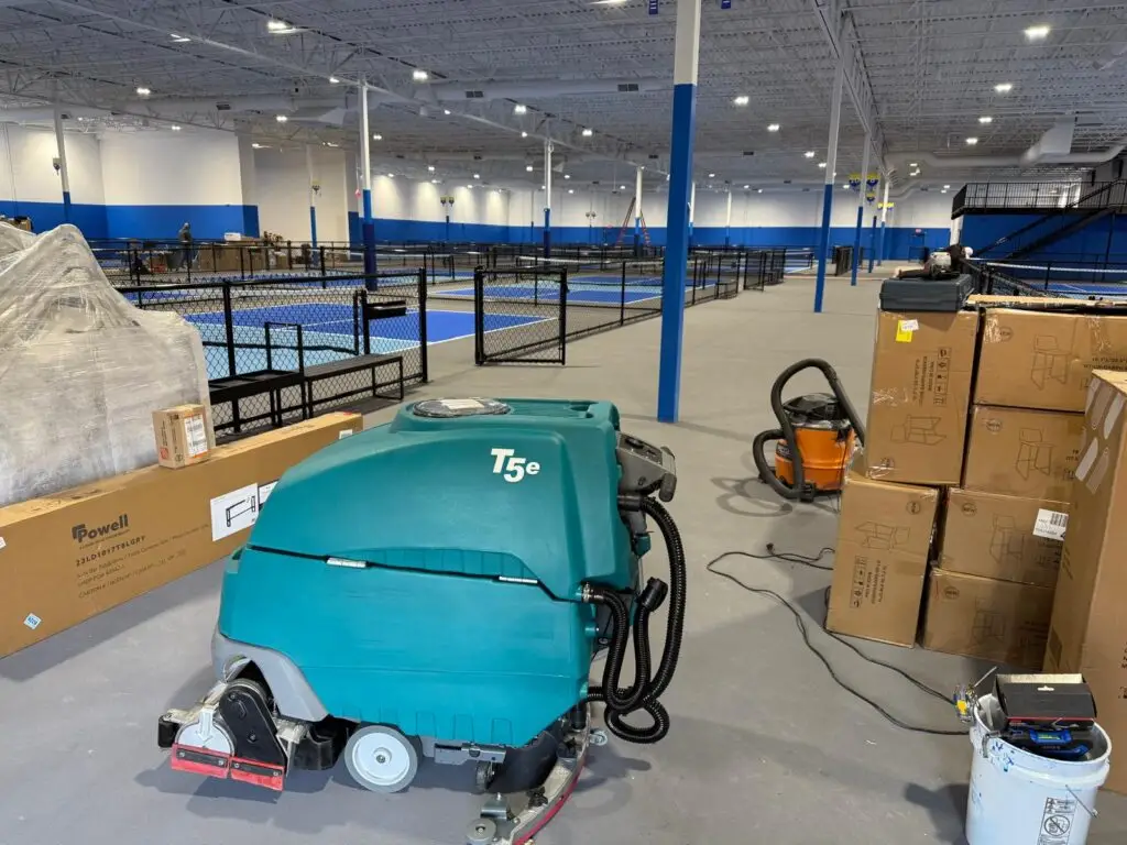 A teal Tennant T5e walk-behind floor scrubber from Robey's PowerKleen parked inside a large indoor sports complex. Cardboard boxes and an orange shop vacuum sit next to the machine, while several blue indoor pickleball courts enclosed by black fencing are visible in the background.