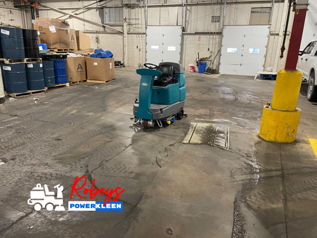 A teal Tennant T7 ride-on floor scrubber from Robey's PowerKleen cleaning a dirty concrete warehouse floor near storage drums and loading dock doors.
