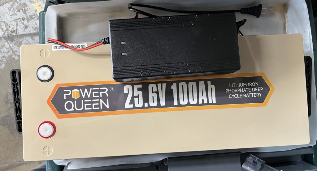 A close-up top view of a Power Queen 25.6V 100Ah LiFePO4 lithium battery with a black power charger resting on top.