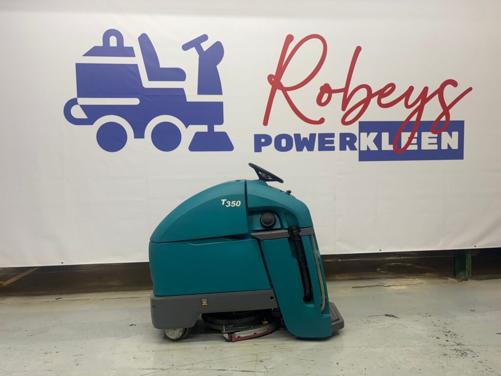 A side profile view of a teal Tennant T350 stand-on commercial floor scrubber from Robey's PowerKleen parked in front of a company backdrop banner.