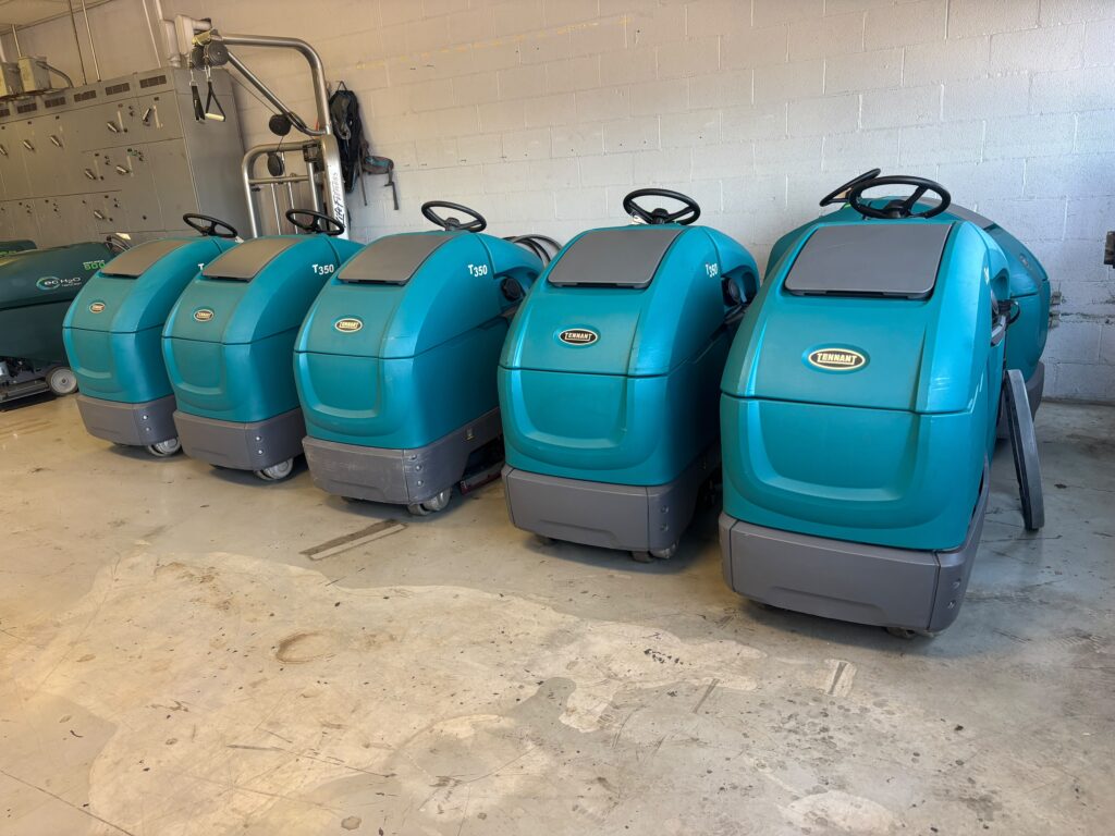 An angled view of five teal Tennant T350 stand-on commercial floor scrubbers lined up in a row at a Robey's PowerKleen equipment facility.