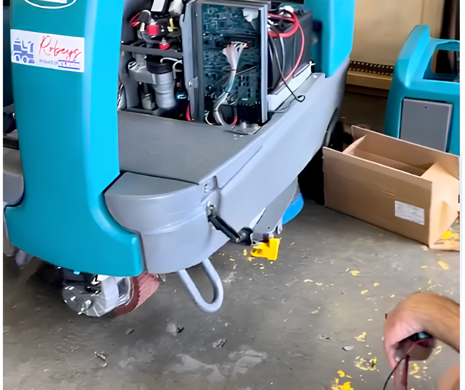 A high-angle view of a floor scrubber machine undergoing maintenance. The main panel is open, exposing a complex arrangement of colored wires and electrical components. A technician’s hands are visible in the foreground, working with a red wire. Part of a turquoise-colored machine with 'Robeys' visible is on the left.